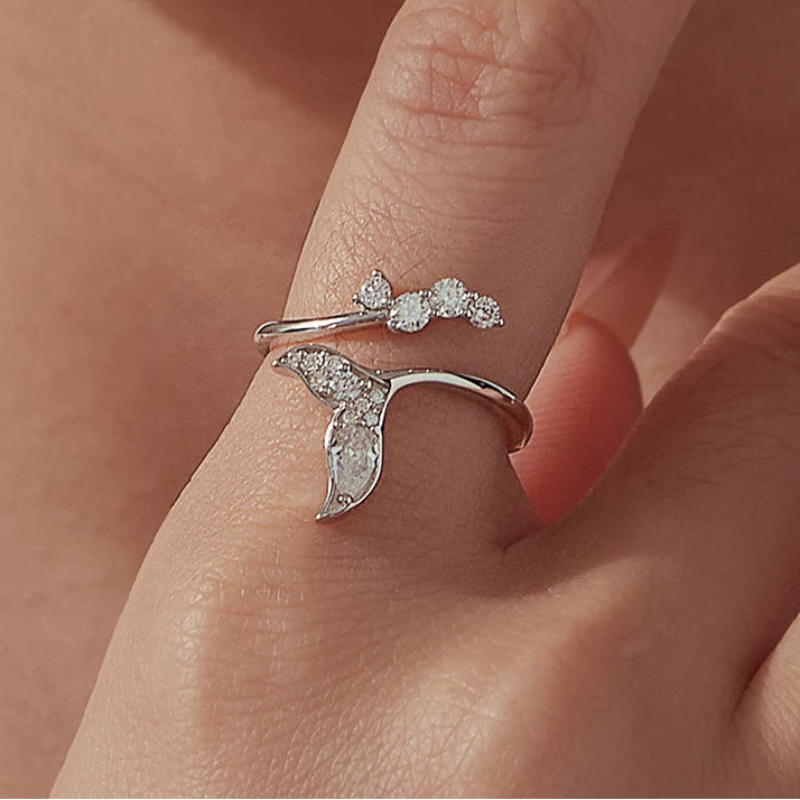 A silver ring with a mermaid tail design and small sparkling stones, shown worn on a finger.