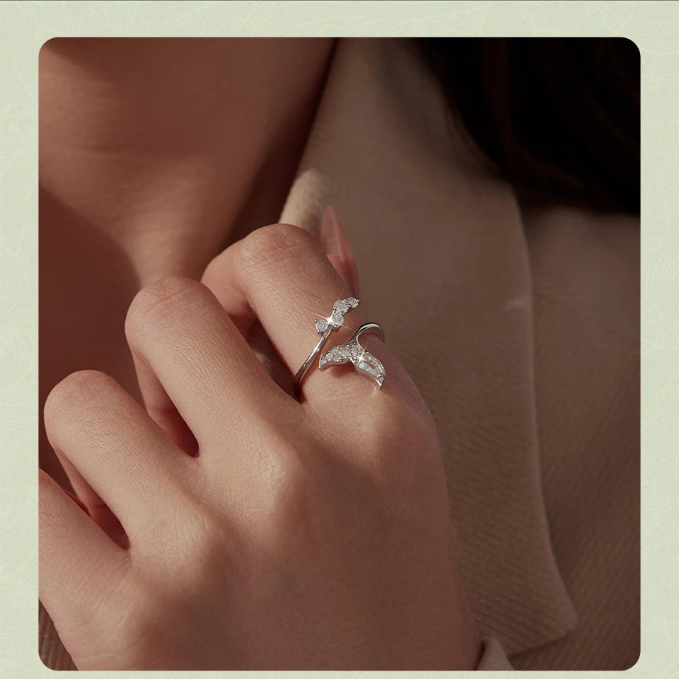 A close-up of a hand wearing two silver bird-shaped rings adorned with small clear stones, set against a soft beige background.