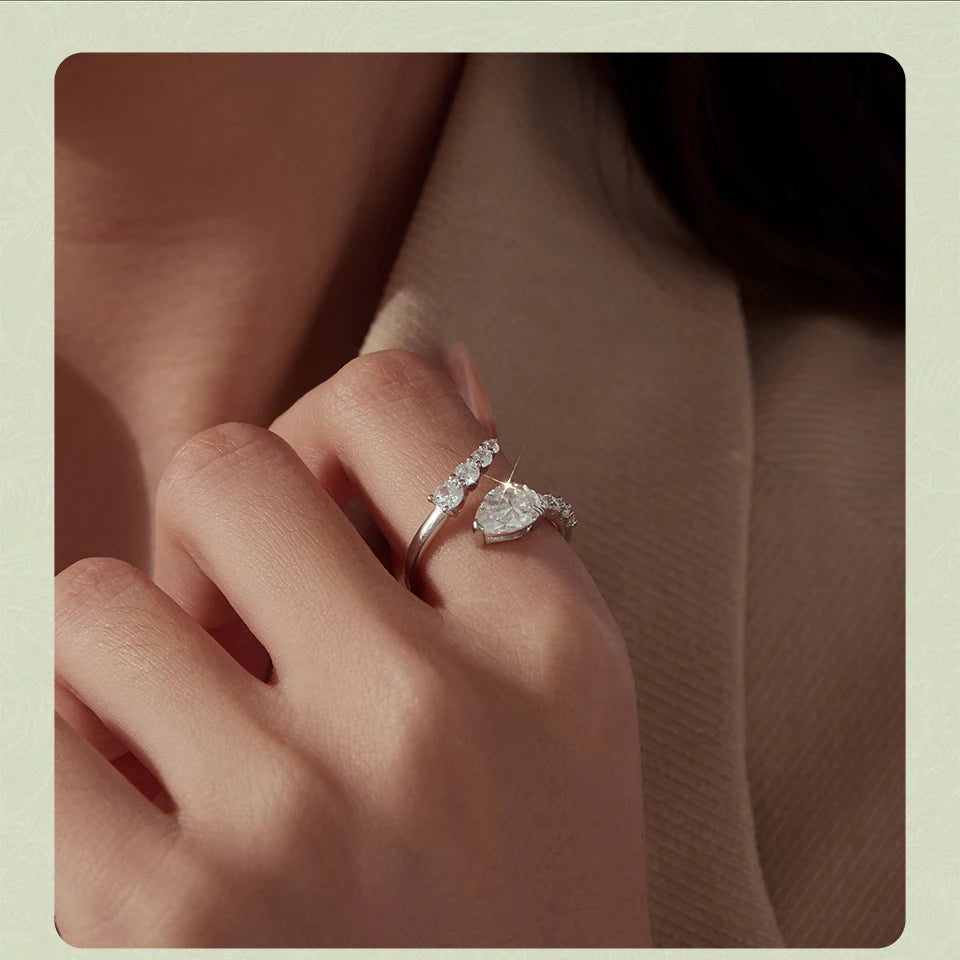 A close-up of a hand wearing two silver rings, including a ring with a heart-shaped clear gemstone and a slim band with small clear stones, set against a soft beige background.