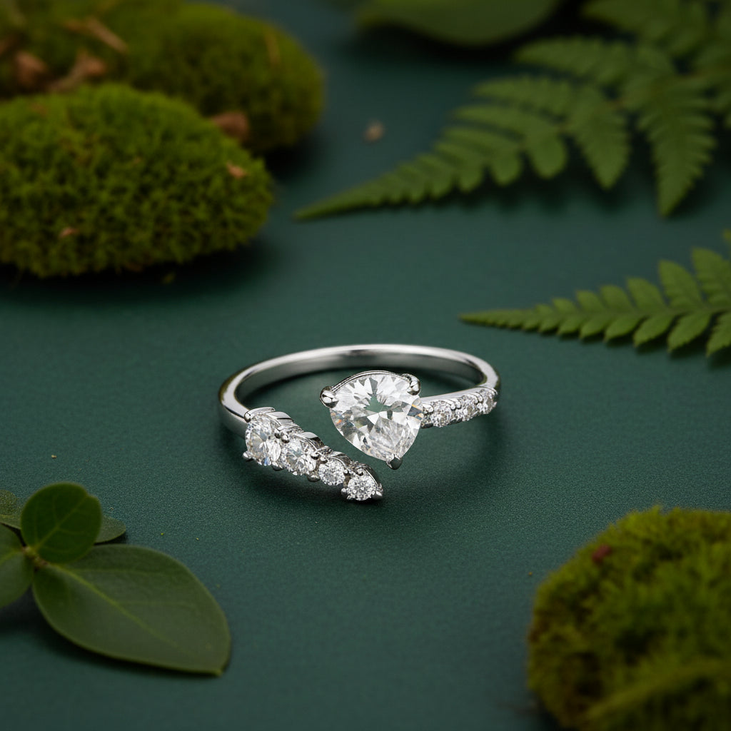 A sterling silver open ring ring with a leaf-like top and a teardrop-shaped clear gemstone, resting on a textured stone with green moss in the foreground.
