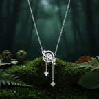 A silver-toned celestial pendant necklace featuring a ring-shaped motif with a central clear gemstone, a small crescent moon, and two dangling star-shaped charms with dark  green woods in the background. 