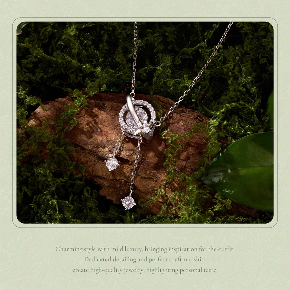 A silver-tone necklace with a circular pavé pendant and two dangling clear gemstone charms, displayed on a mossy log with decorative text below.