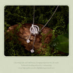 A silver-tone necklace with a circular pavé pendant and two dangling clear gemstone charms, displayed on a mossy log with decorative text below.
