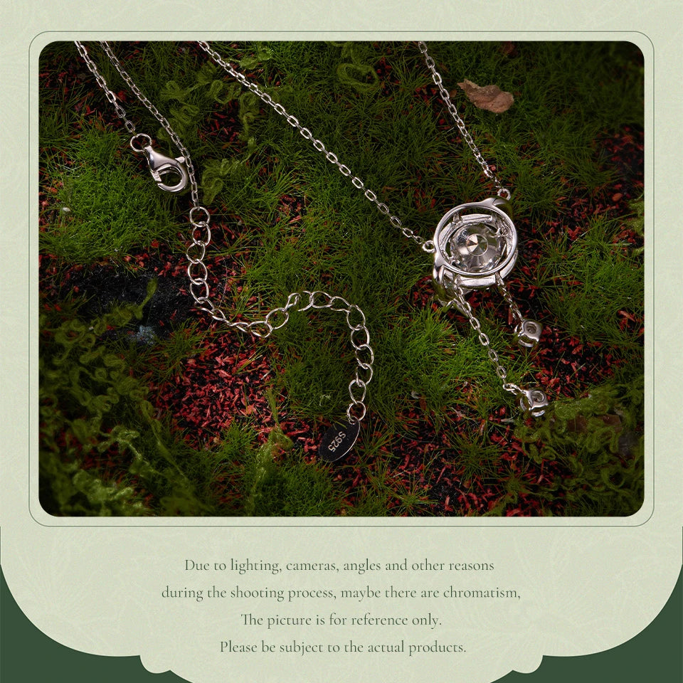 A silver-toned chain necklace with a round clear stone pendant and small dangling beads, laid on dark mossy ground. The photo is framed by a pale green border with disclaimer text below.