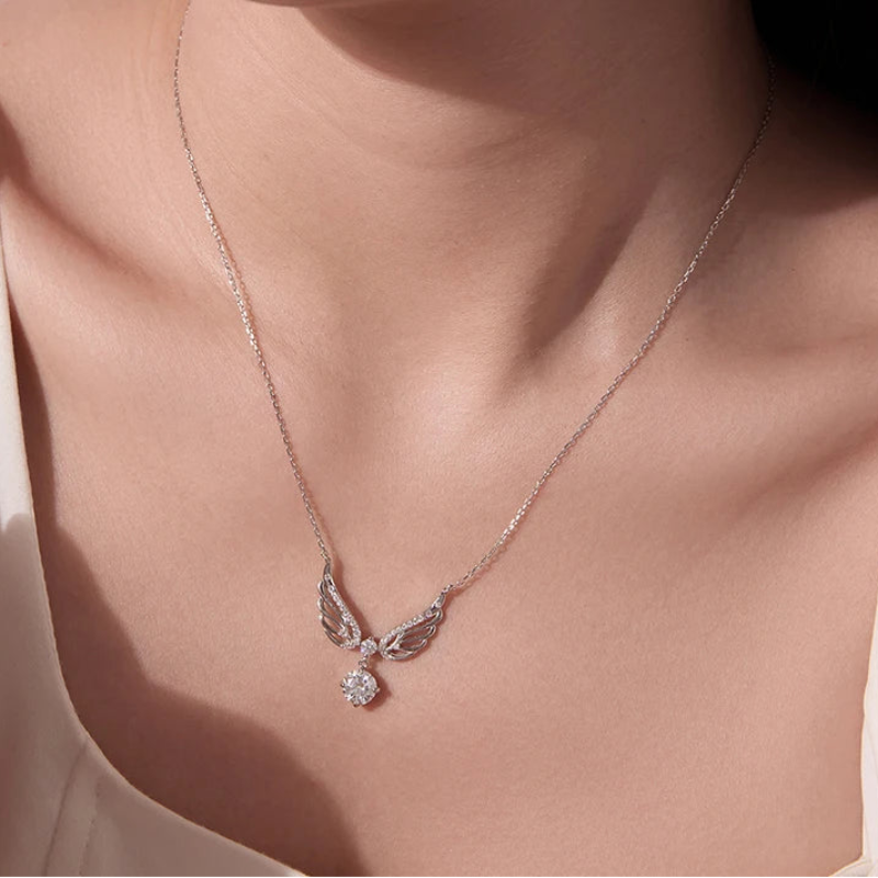 A close-up of a model wearing a delicate silver chain necklace with a winged pendant and a small clear gemstone, against a warm beige top.
