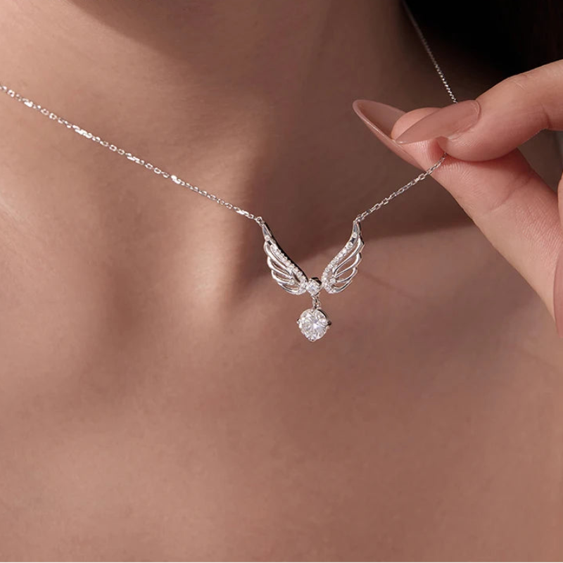 A close-up lifestyle shot of a model wearing a silver angel wing pendant necklace with a clear faceted gemstone, with a hand gently holding the chain.