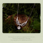 A silver-toned angel wing pendant necklace with a clear round gemstone, displayed on a mossy brown log with green leaves. The photo is framed with a light green border containing descriptive text at the bottom.