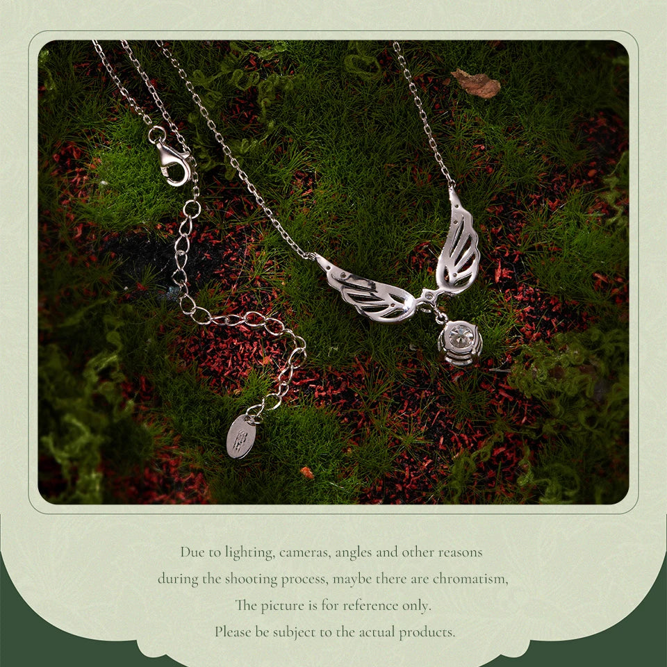 A silver-toned angel wing pendant necklace with a small rose charm and a separate oval tag charm, displayed on green moss with red leaves. The photo is framed by a light green border with disclaimer text below.