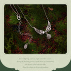 A silver-toned angel wing pendant necklace with a small rose charm and a separate oval tag charm, displayed on green moss with red leaves. The photo is framed by a light green border with disclaimer text below.