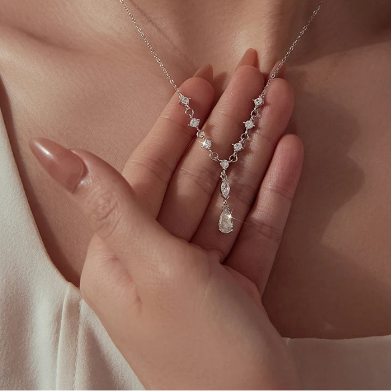 A close-up of a model wearing a delicate silver necklace with clear teardrop-shaped crystals and small star-shaped clear stones. The model's hand is resting on her chest.