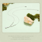 A delicate silver necklace with a small cluster of clear stones at the center, displayed on a pale mint green background with decorative moss and a stone. Chinese promotional text is printed at the bottom.