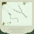 A silver-toned delicate necklace with a small teardrop-shaped pendant and an adjustable chain extender, displayed on a pale mint background with decorative moss and a note of uncertainty about lighting and color accuracy.