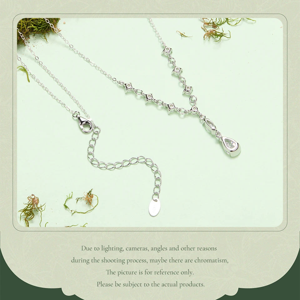 A silver-toned delicate necklace with a small teardrop-shaped pendant and an adjustable chain extender, displayed on a pale mint background with decorative moss and a note of uncertainty about lighting and color accuracy.