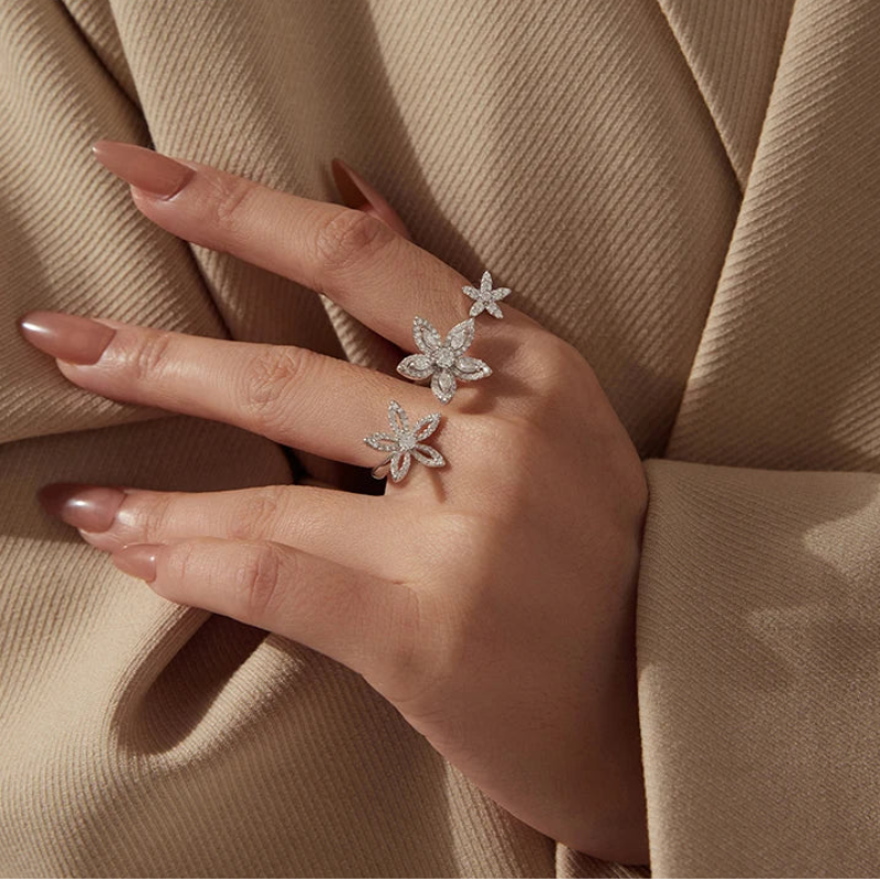 A close-up of a hand wearing three silver flower-shaped rings set with clear stones, resting against a beige textured fabric background.