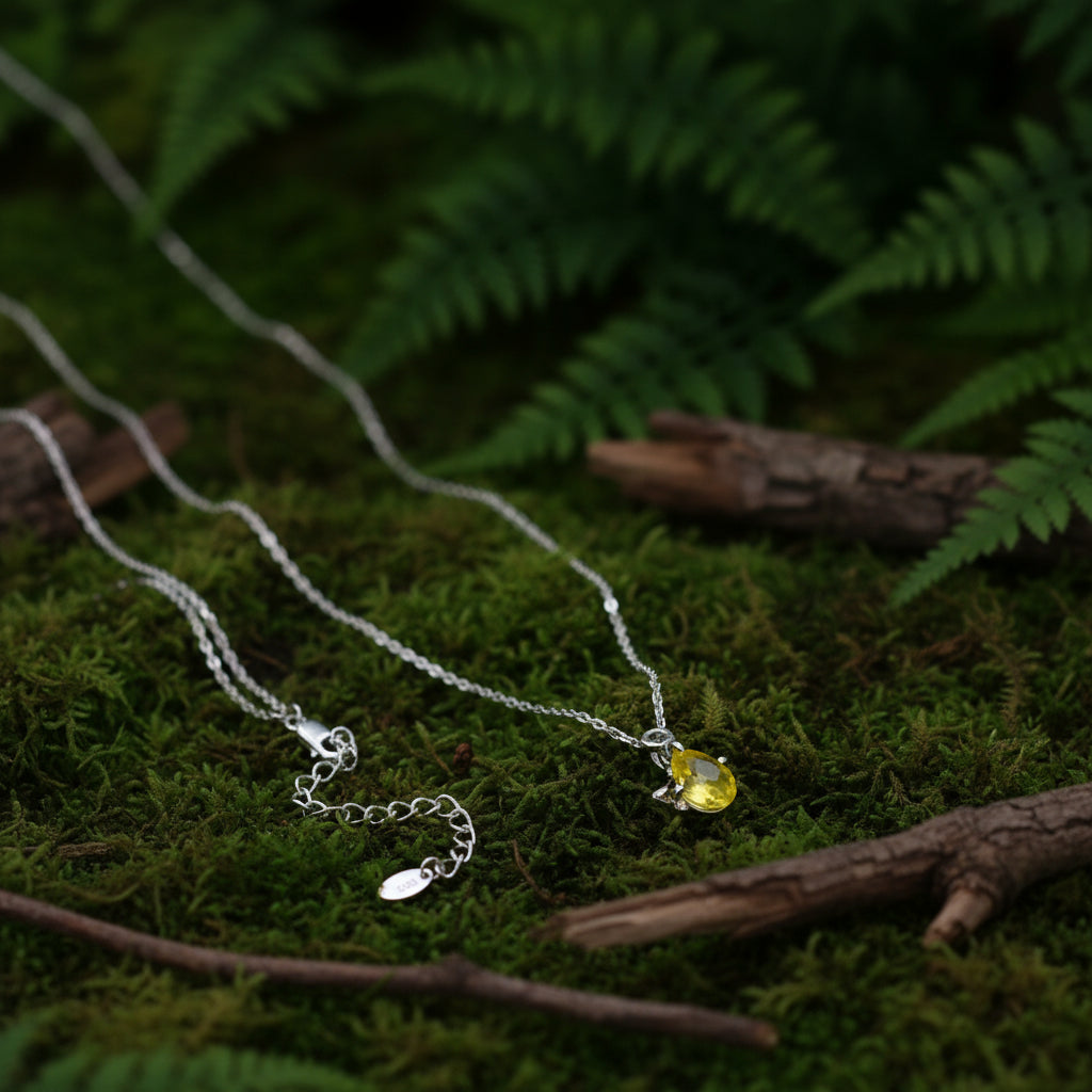 A silver chain necklace with an adjustable lobster clasp extension chain and a small oval tag, displayed alongside a matching pendant featuring a yellow-green faceted gemstone, set against a grass in the background.