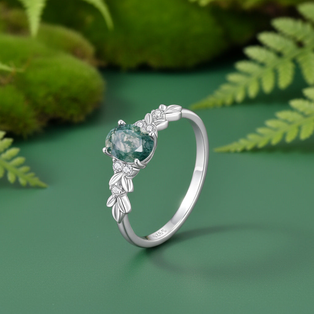 A silver ring with a oval-shaped green gemstone and delicate leaf-shaped accents, shown on a green background.