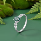 A silver ring with a oval-shaped green gemstone and delicate leaf-shaped accents, shown on a green background.