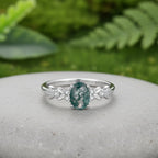 A silver ring featuring an oval green marbled gemstone in the center, flanked by small clear accent stones and leaf-shaped silver details on a grey stone background.