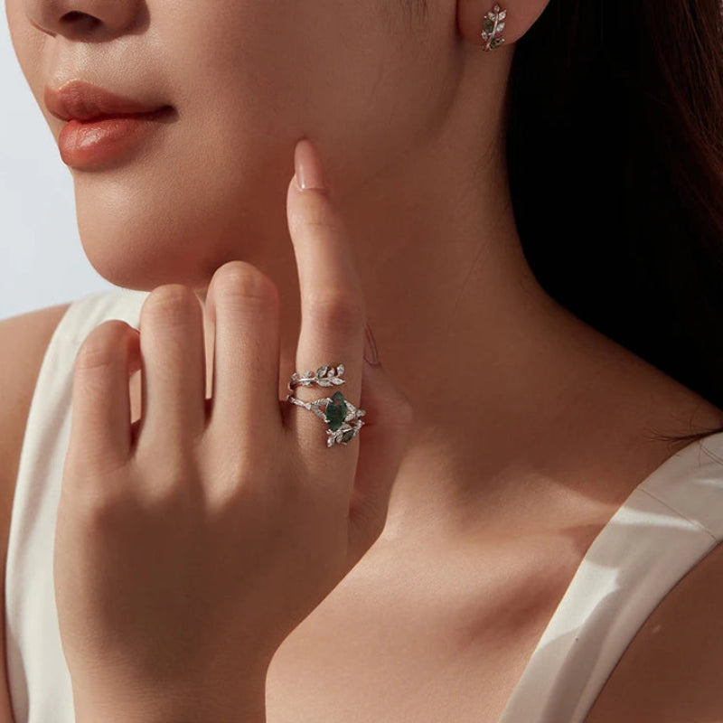 A close-up lifestyle shot of a model wearing a silver ring with a green rectangular gemstone and small clear stones, along with a matching earring.