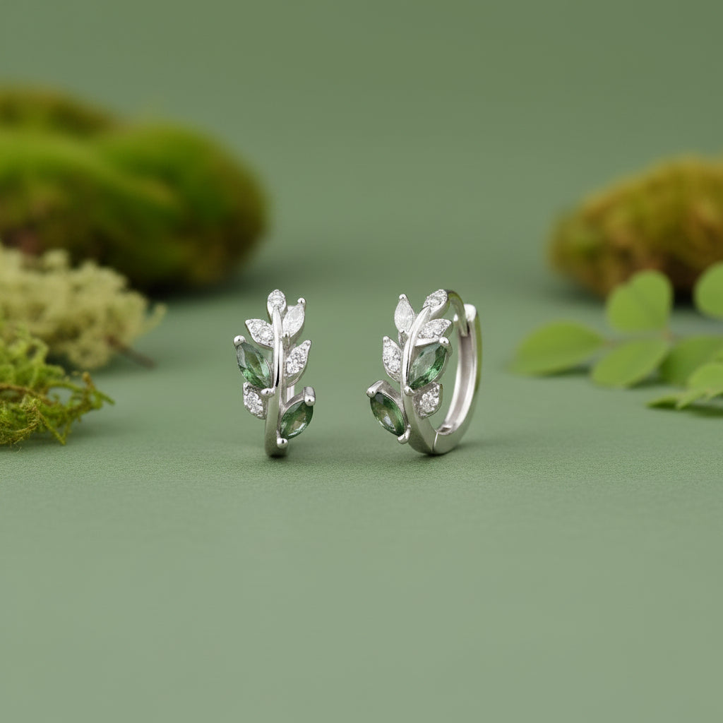 Silver huggie hoop earrings with green gemstone accents and clear crystals, displayed on a piece of sage green table against a soft green background.