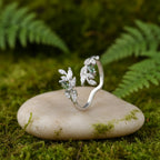 A silver-toned ring with leaf-shaped crystal clusters and a small grey stone, displayed on a piece of stone against a soft green background.
