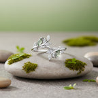 A silver branch ring with marquise-cut green gemstones and small clear stones, shown on a sage green background.