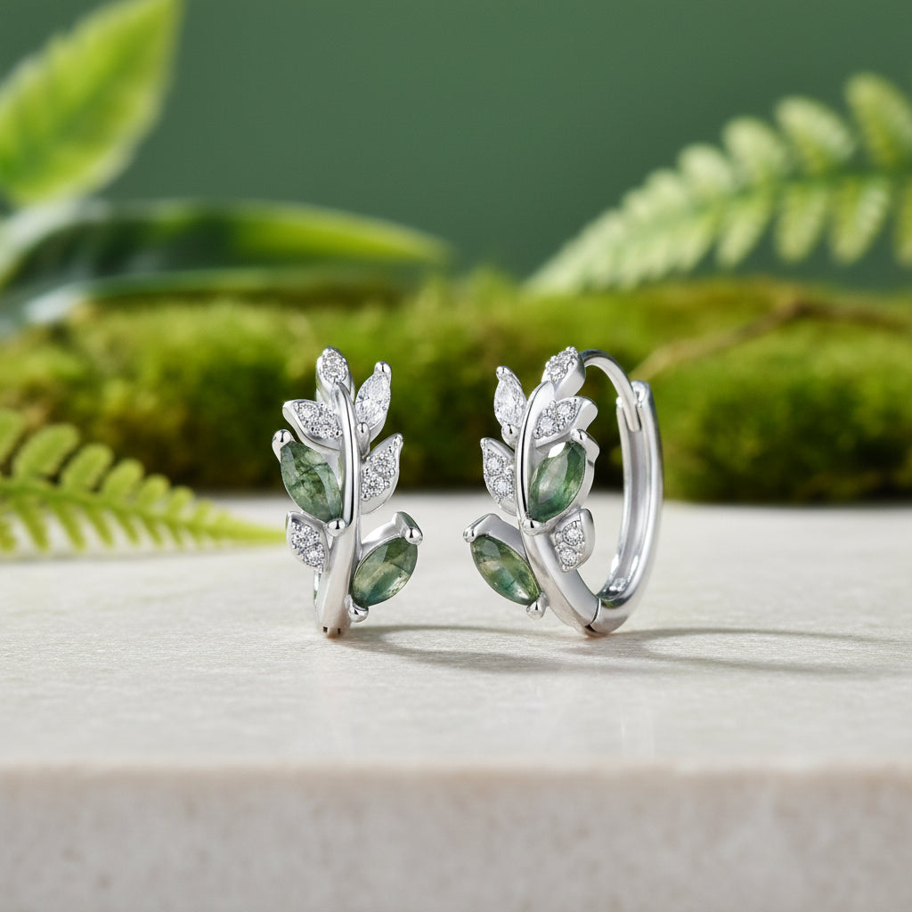 Silver huggie hoop earrings with marquise-cut green gemstones and clear accent stones displayed on the white marble, sage green and leaves in the background.