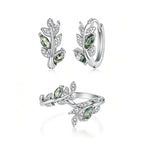 A jewelry set featuring a silver huggie hoop earring, a matching leaf-shaped ring, all adorned with green marquise-cut gemstones and clear pavé crystals on a white background.