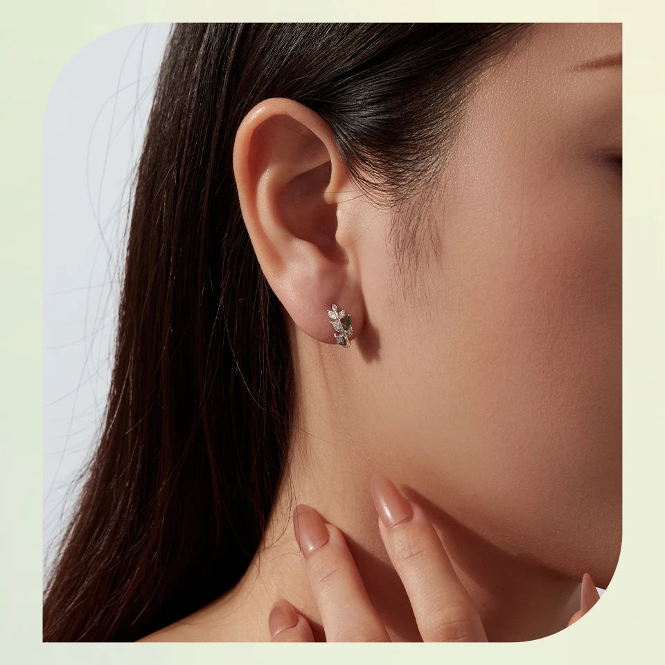 A close-up of a woman's ear wearing a small silver-toned stud earring with clear stones, shown against a soft cream background.