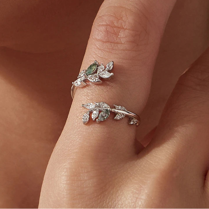 A close-up of a hand wearing two silver leaf-shaped rings with green and clear stones.