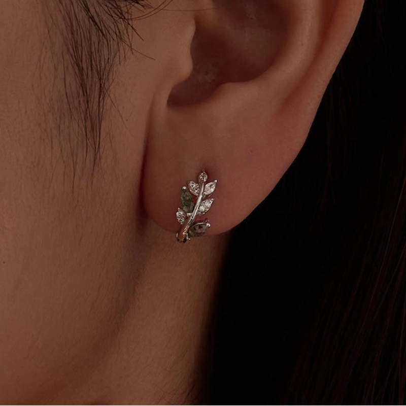 A close-up of a person's ear wearing a small leaf-shaped stud earring with clear stones and green accents.