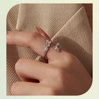 A close-up of a hand wearing two delicate silver-toned rings with floral and leaf motifs set with clear stones, resting against a beige textured fabric background.