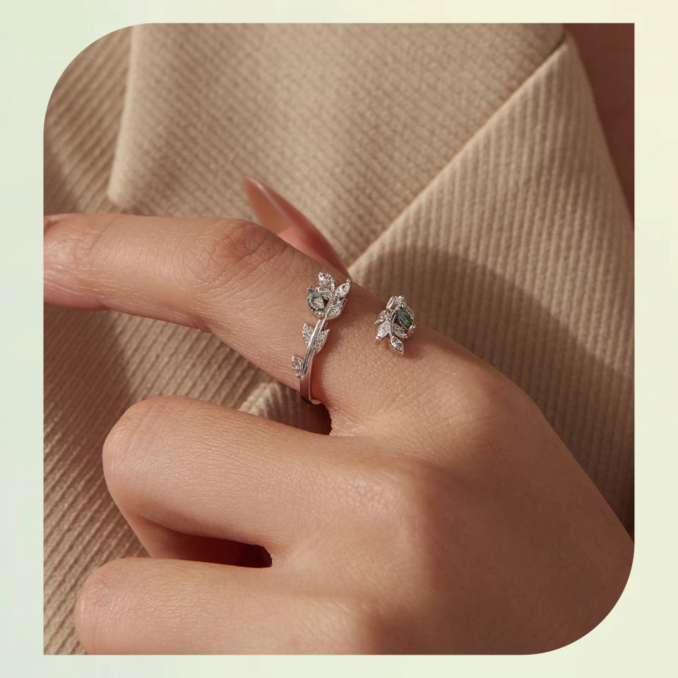 A close-up of a hand wearing two delicate silver-toned rings with floral and leaf motifs set with clear stones, resting against a beige textured fabric background.