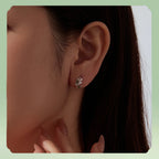 A close-up of a woman's ear wearing a small silver leaf-shaped stud earring with clear stones, set against a soft light green background.