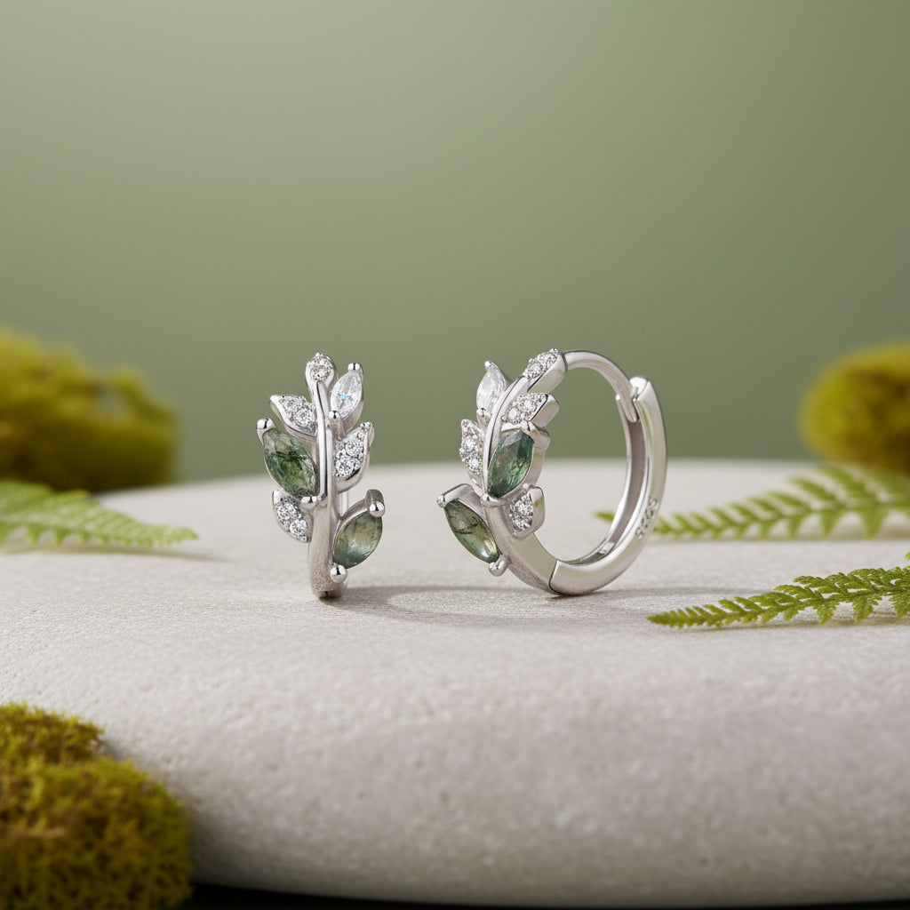 A pair of sterling silver huggie hoop earrings with leaf-shaped green gemstone accents and clear crystal details, displayed on a piece of stone against a soft green background.